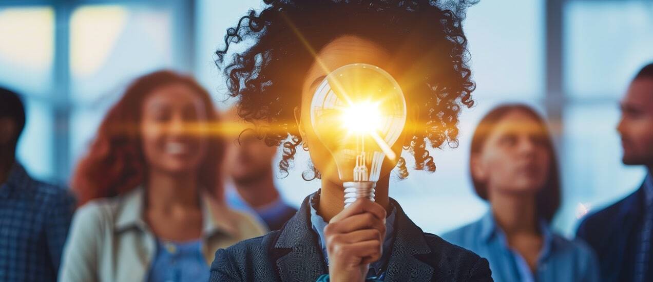 Individual holding a lighted bulb in front, concept of idea creation with peers in background