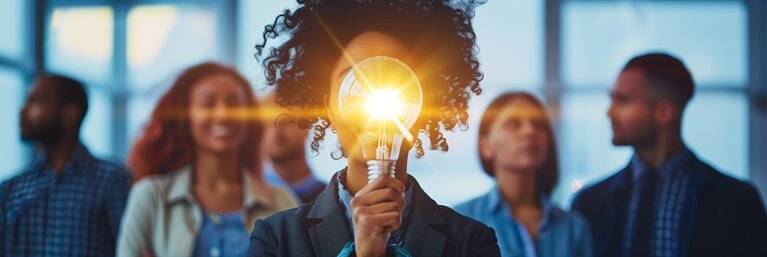 Individual holding a lighted bulb in front, concept of idea creation with peers in background